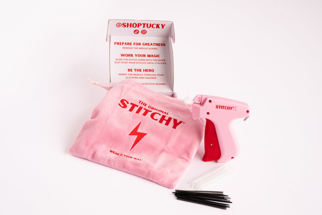 A pink fabric pouch printed with the Stitchy name, beside its pink fastening tool and a row of black fastener pins, in front of the Stitchy packaging.